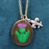 An oval pendant of resin covering a purple thistle with green foliage, hanging from a brass chain